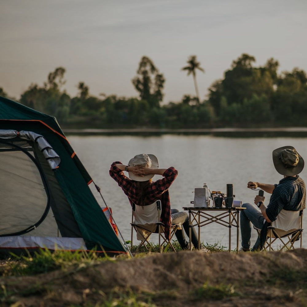 Two people camping by a lake, sitting at a small table with drinks and a laptop.