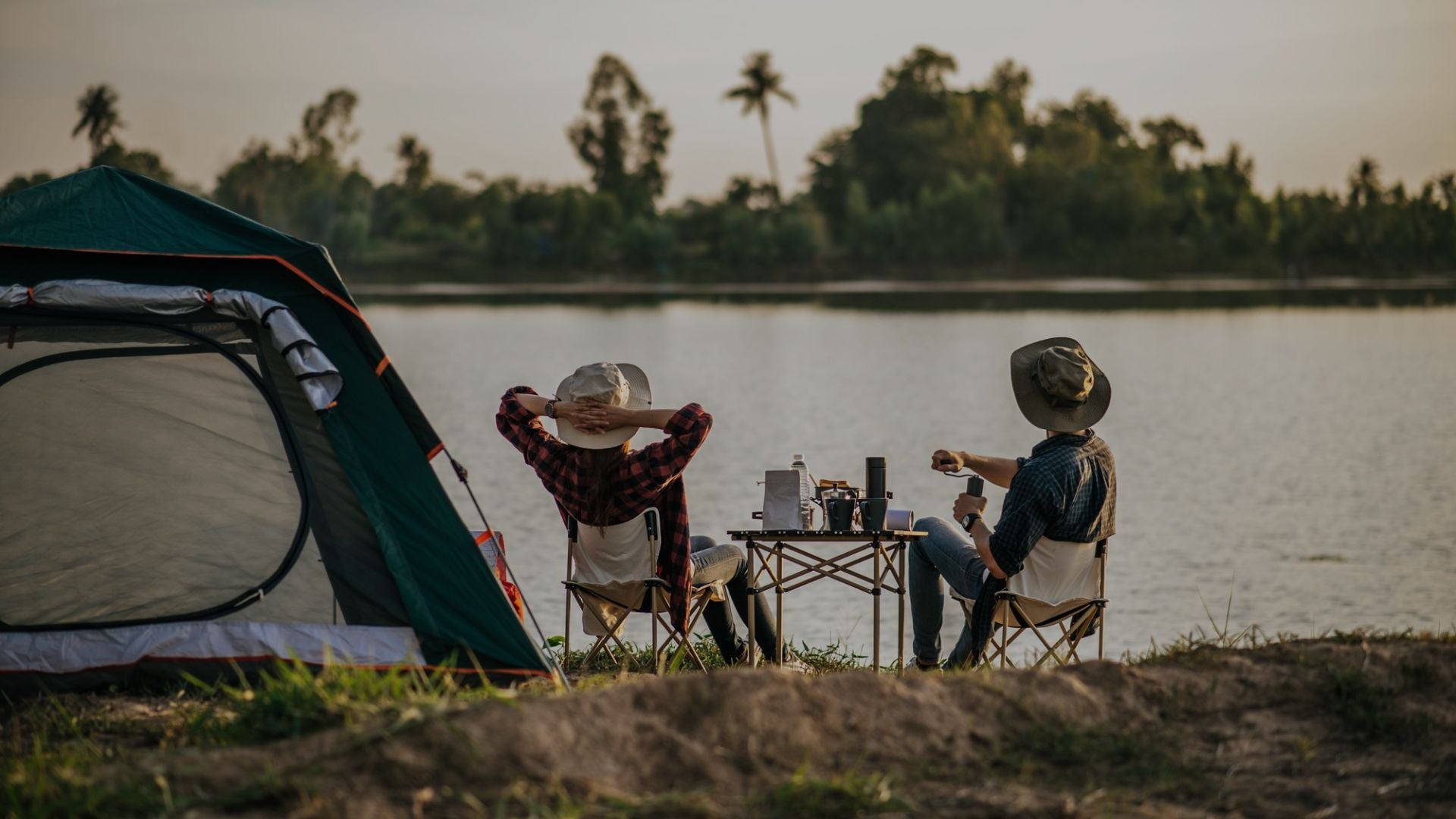 Two people camping by a lake, sitting at a table with drinks and a tent nearby.
