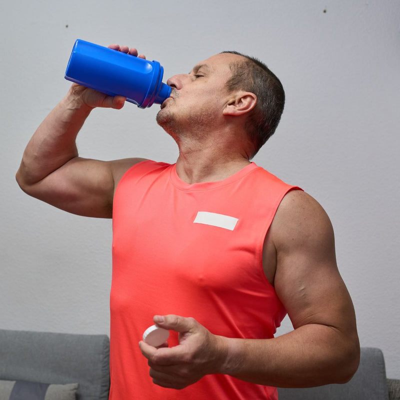 Man in a red tank top drinking from a blue shaker bottle against a gray background